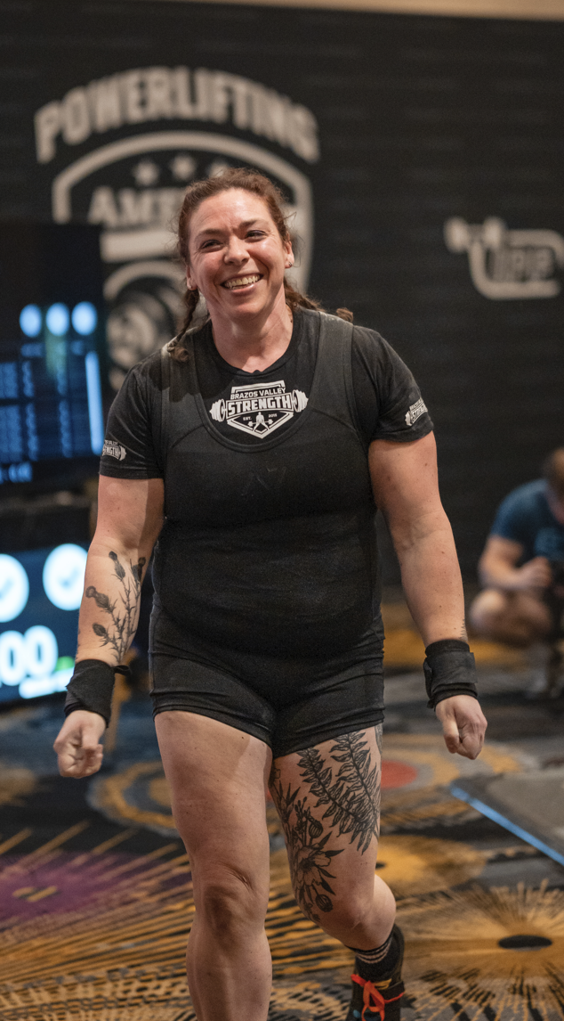 Woman in athletic wear with tattoos, standing on a powerlifting platform.